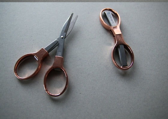 Grey background with rose gold fold away scossors shown in two positions L-R: fulley extended and blades closed, fully closed.