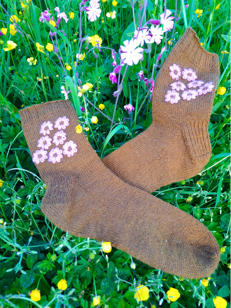 Close up of  Blathan hand knitted Socks in a khaki green with pink embroidered flowers as a flatlay in a meadow