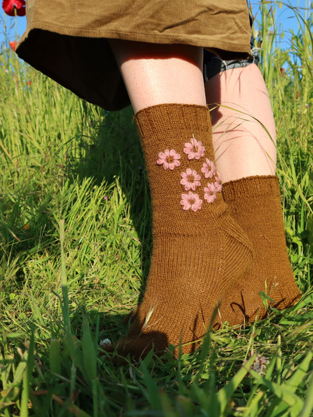Close up Blathan hand knitted Socks being worn in a meadow in a khaki green with pink embroidered flowers.