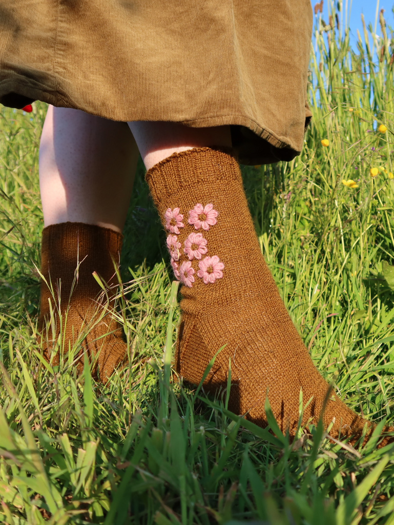Stood in a meadow wearing Blathan hand knitted Socks in a khaki green with pink embroidered flowers 