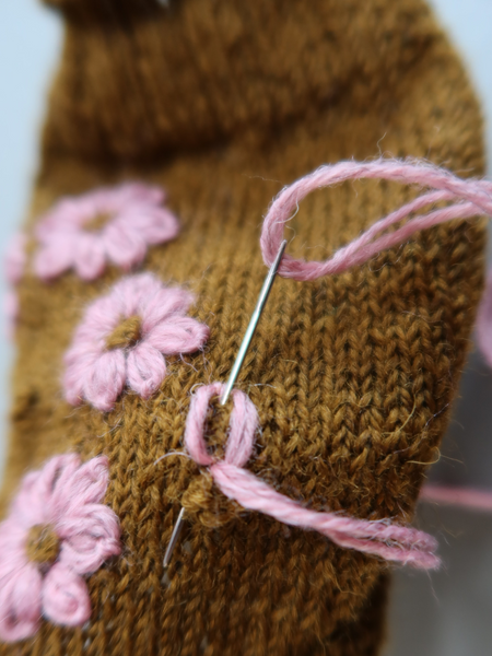 Close up of  Blathan hand knitted Socks in a khaki green with pink embroidered flowers being made with a needle