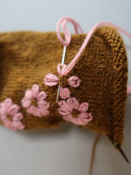 Close up of  Blathan hand knitted Socks in a khaki green with pink embroidered flowers being made with a needle