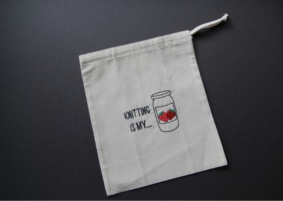 Grey background with natural cotton bag embroidered with "Knitting is my..." and a jar of strawberry jam beside the text.