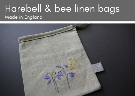 Grey background with natural linen bag, embroidered with purple harebells, a yellow sprig of grass and a bumple bee.