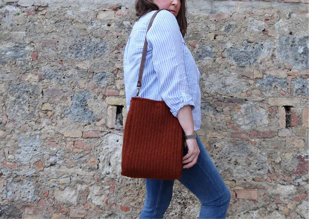 Rust coloured crocheted bag in rust aran weight wool over shoulder of woman beside an old stone wall.