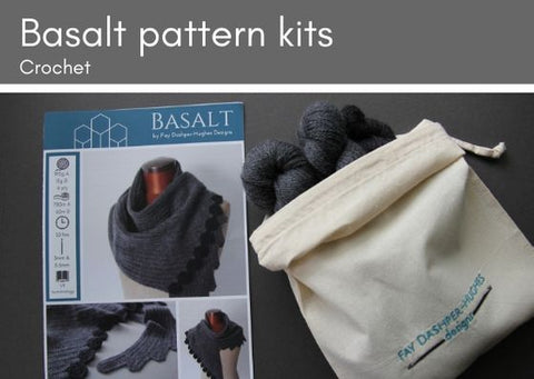 Basalt crocheted shawl kit: on the left lies a hardcopy of the pattern showing the shawl in two shades of grey, the darkest is used for the hexagonal edging.  To the right is a folded down bag with embroidery saying "Fay Dashper-Hughes Designs".  Inside the bag are two large skeins of a mid-grey Merino wool and a mini skein of a charcoal grey Merino wool.