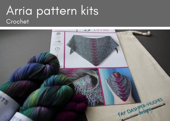 Arria crocheted shawl kit: two skeins of variegated wool (pinks, purples, greens, teals and blues) lie on top of a pattern for the shawl which is on top ofa cotton bag with "Fay Dashper-Hughes Designs" embroidered on it.