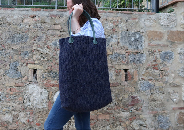 Dark denim blue crocheted bag in  chunky weight wool held up by a woman beside an old stone wall.