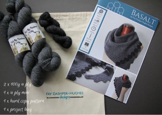 Basalt crocheted shawl kit:  In the centre is a cotton bag with embroidery saying "Fay Dashper-Hughes Designs".  On top of the bag are two large skeins of a mid-grey Merino wool and a mini skein of a charcoal grey Merino wool.   On the right lies a hardcopy of the pattern showing the shawl in two shades of grey, the darkest is used for the hexagonal edging. 